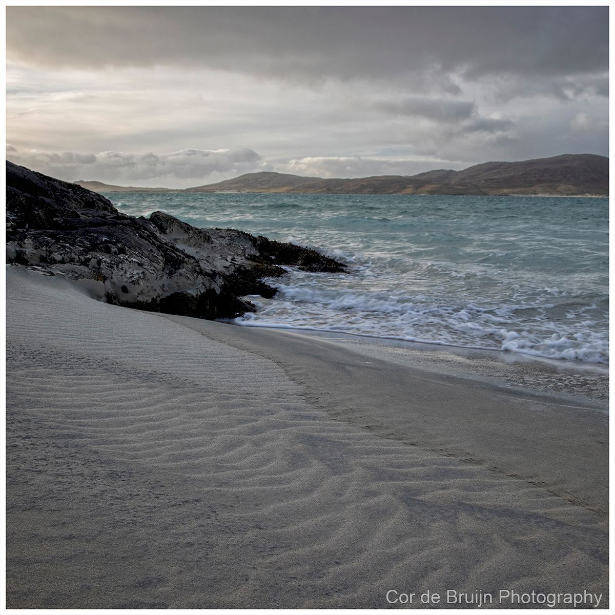 Sandy beach with rippled textures, waves, rocks, and hills under a cloudy sky.