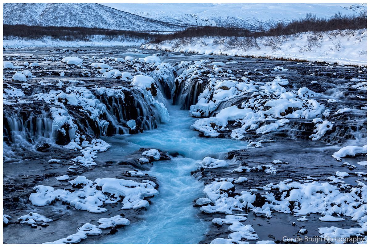 Frozen waterfall with blue water flowing through ice and snow-covered rocks, mountains in the distance.