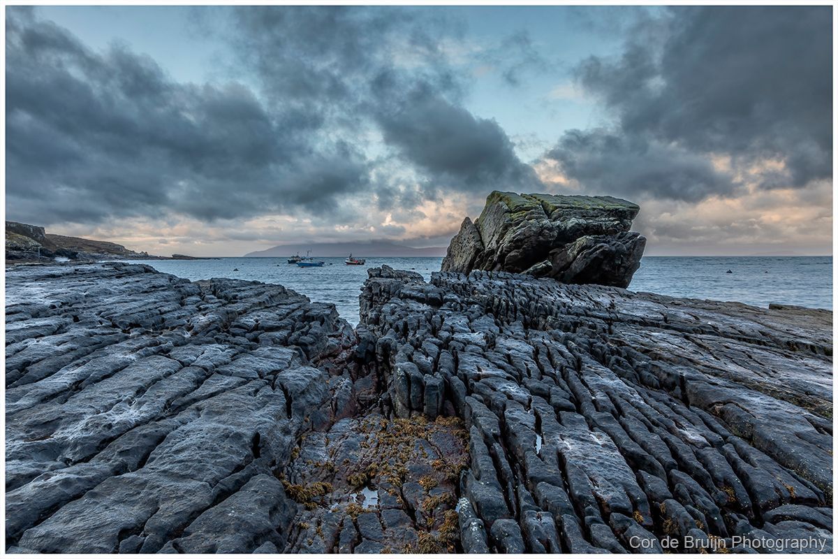 Rocky shoreline at dusk with a large boulder under a cloudy blue-gray sky.