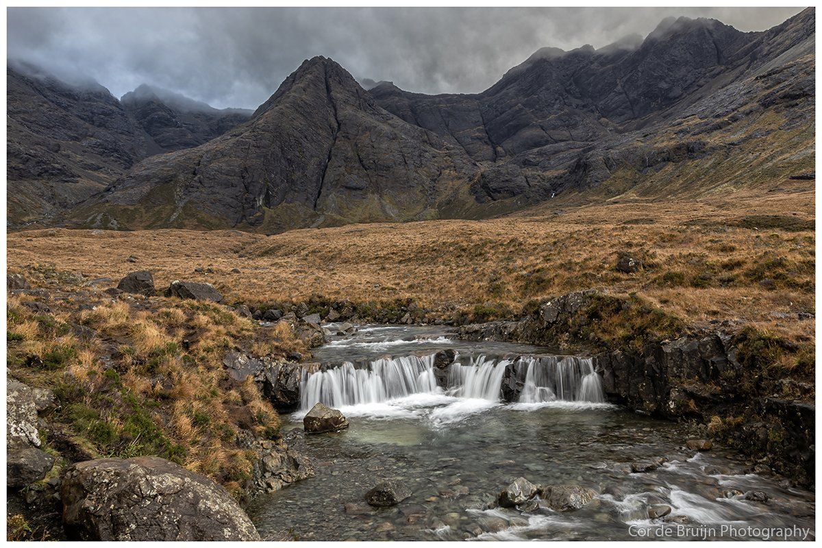 Rocky mountain landscape with a small waterfall flowing through a grassy plain under a cloudy sky.