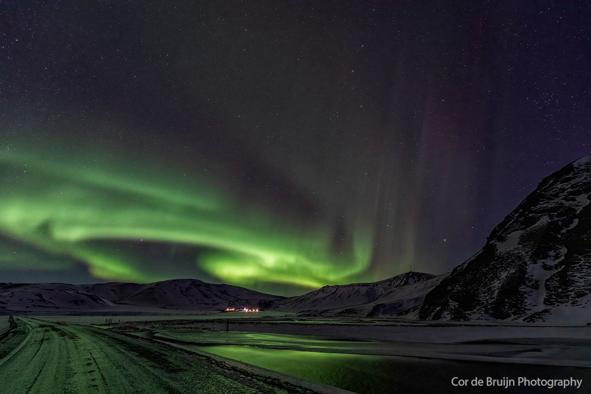 Green aurora borealis swirls across a starry night sky over snowy mountains and a road.