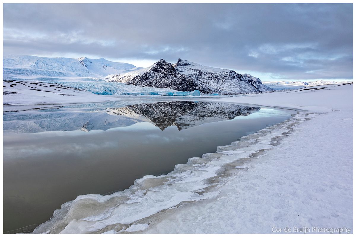 Snowy mountain landscape reflected in a calm icy lake under a cloudy sky