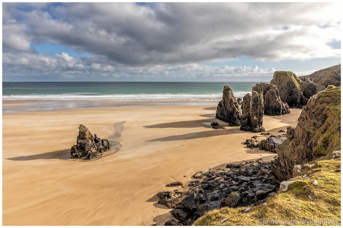 Sandy beach with rock formations under a cloudy sky. Ocean waves in the distance.