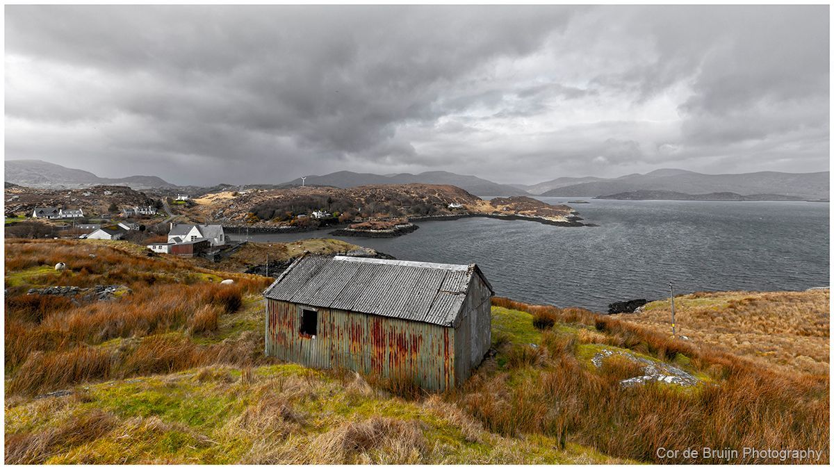 Dilapidated corrugated metal shack overlooking a dark, stormy sea and a distant town under a cloudy sky.