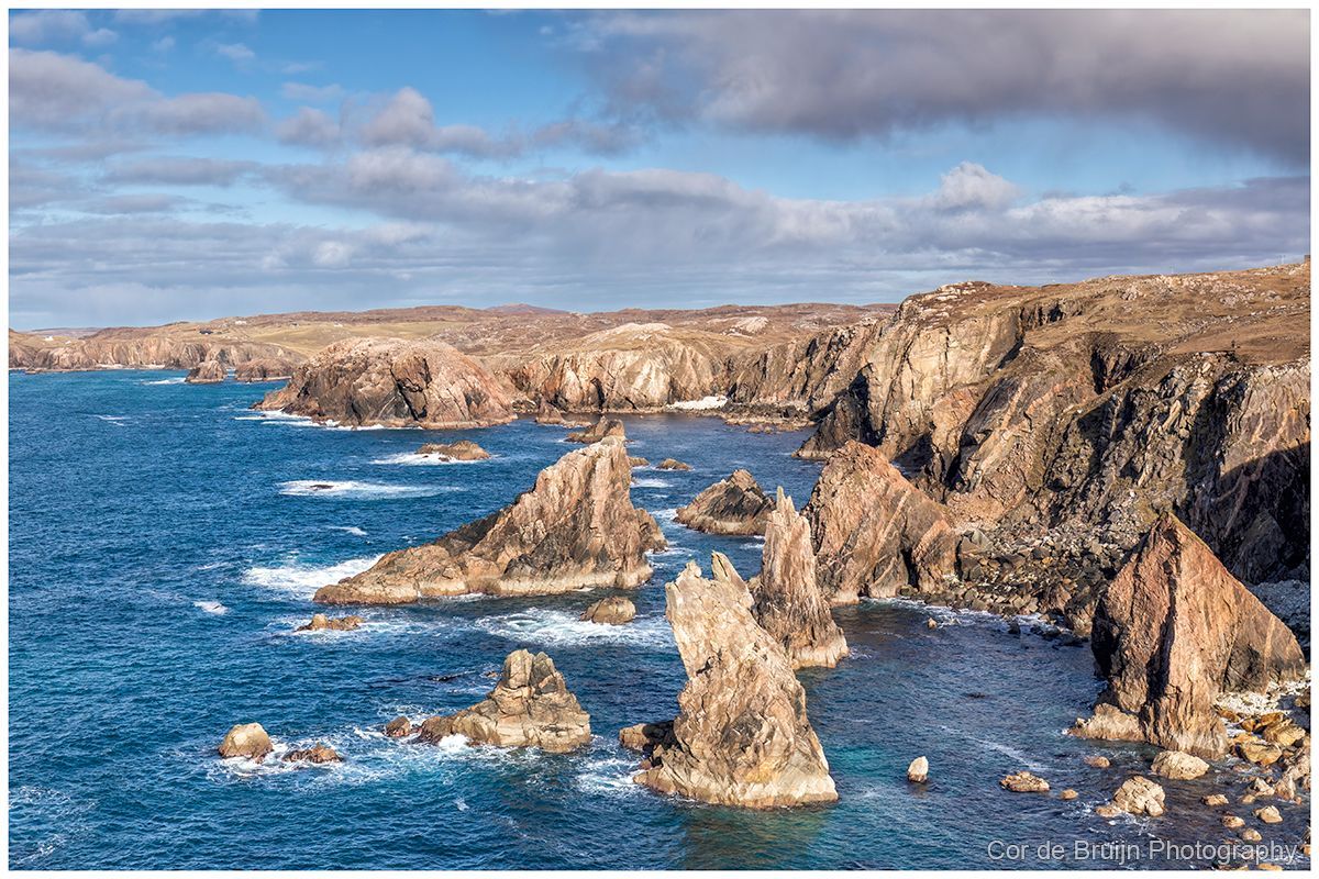 Rocky coastline with jagged rock formations jutting from a deep blue ocean under a partly cloudy sky.
