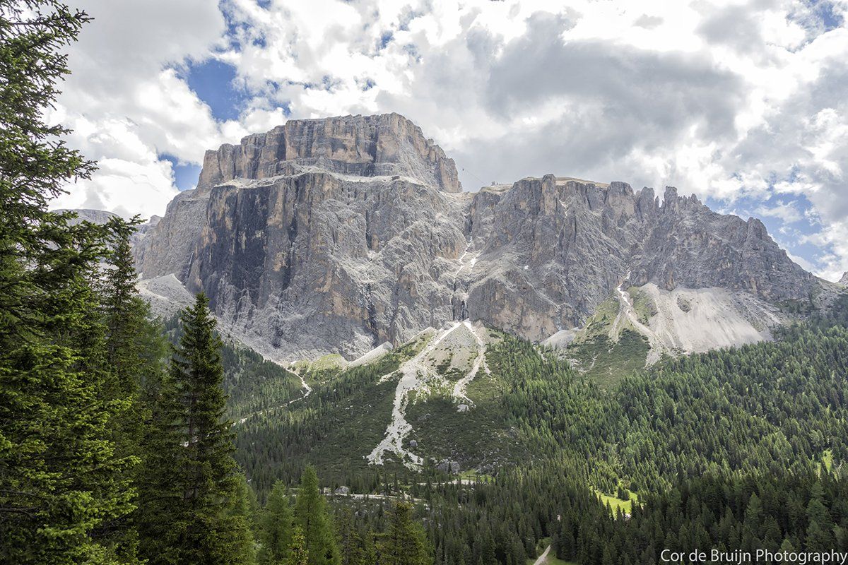 A towering, rocky mountain with a forested foreground under a cloudy sky.