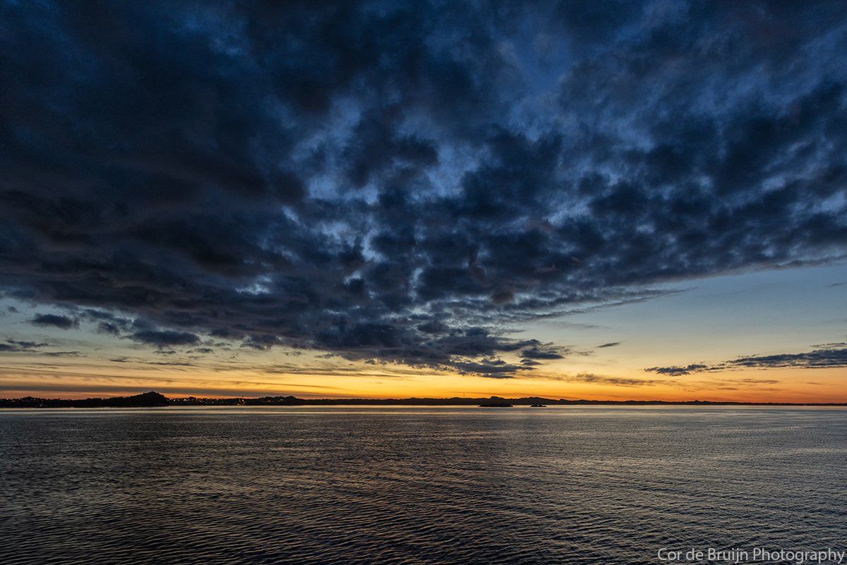 Dramatic sky at dusk over rippled water. Orange and yellow glow on the horizon, dark clouds above, silhouette of land.