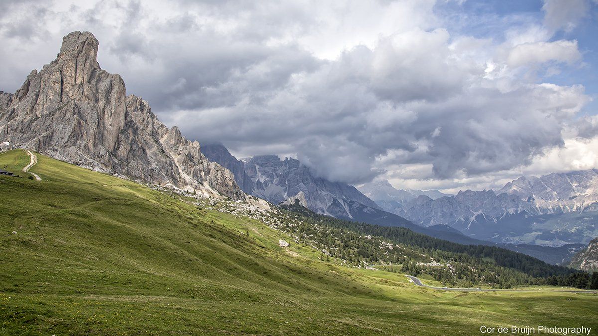 Green hillside leads to rugged mountain range under cloudy sky.