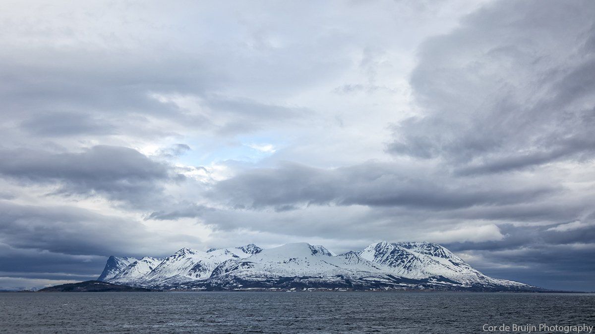 Snow-capped mountain range under a cloudy sky, reflected in the still water below.