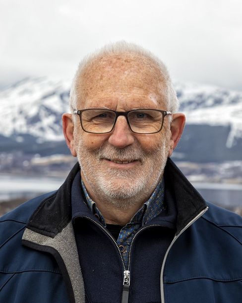 Man wearing glasses, smiling, with mountains and water in the background.