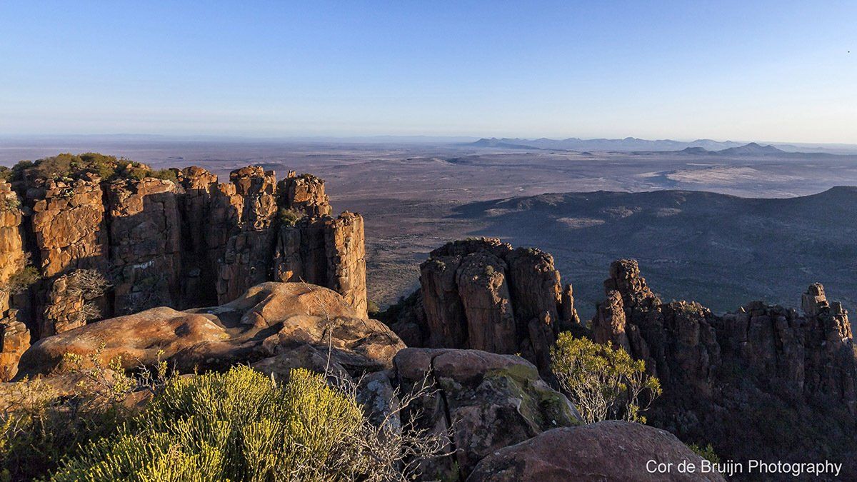 Rocky mountain ridge overlooking a vast, arid landscape under a clear blue sky.