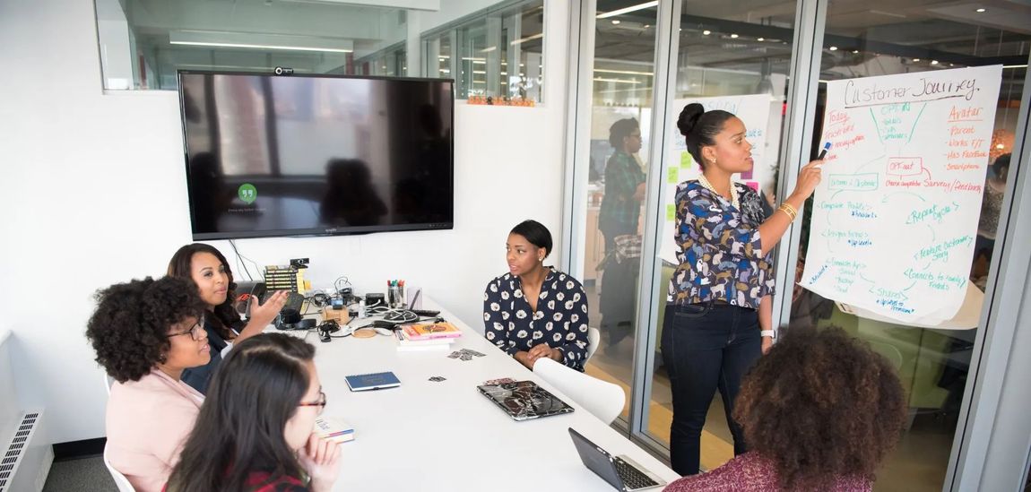 Group of women in a meeting, one presenting at a whiteboard with notes.
