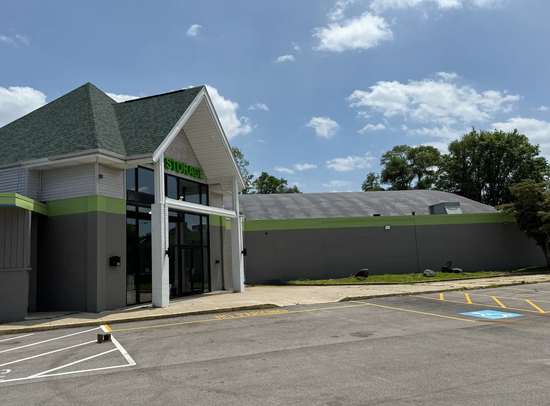 Empty storefront with green trim beside a parking lot under a partly cloudy sky