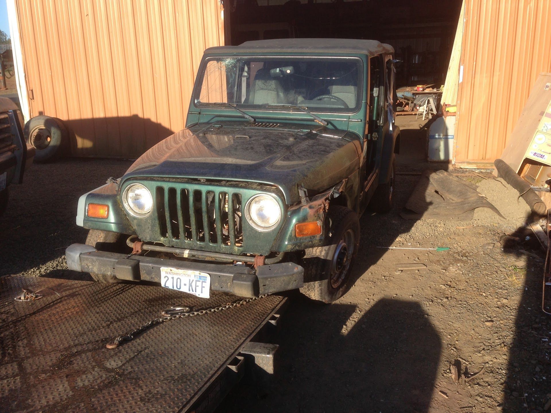 Green Jeep Wrangler on a flatbed tow truck in front of a tan barn. The Jeep's front end is damaged | South Beach Towing & Repair