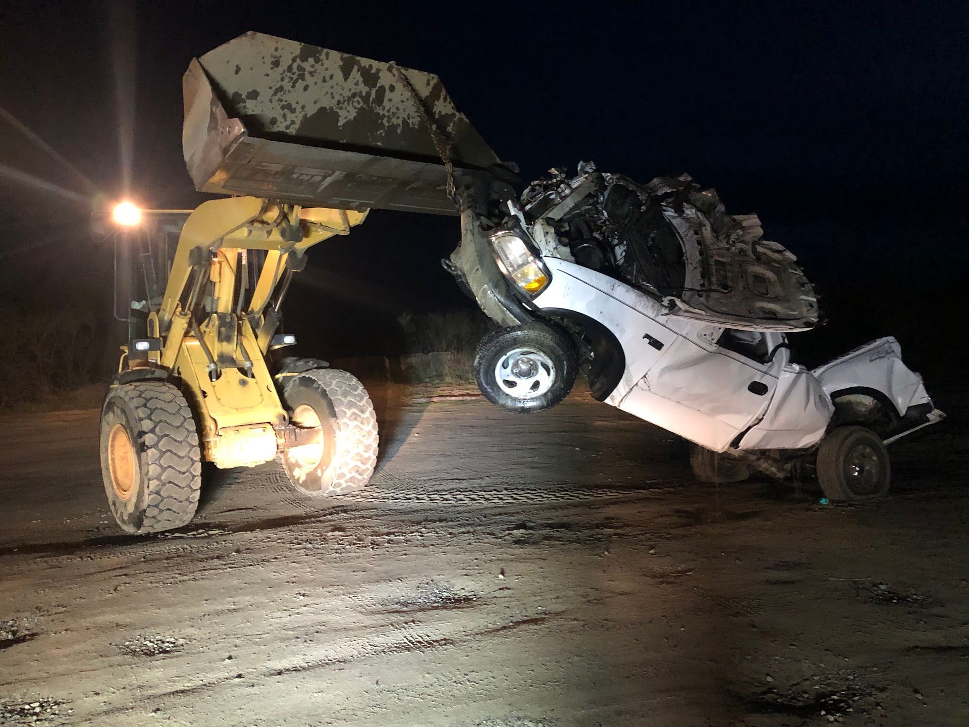 A damaged white pickup truck being lifted by a yellow front-end loader at night | South Beach Towing & Repair