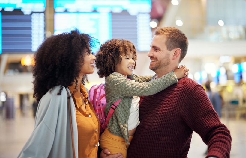 Family embracing in an airport terminal, smiling near blurred departure boards.