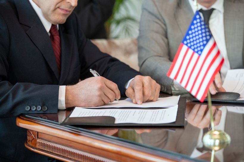 Businesspeople signing documents at a desk beside a small American flag
