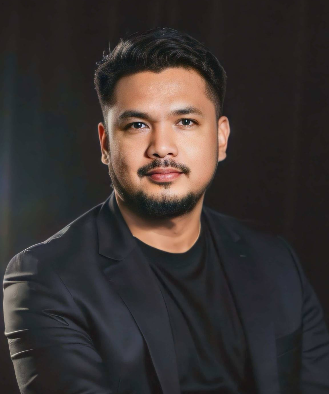 Studio portrait of a person in a black blazer and shirt, seated against a dark background with a calm expression