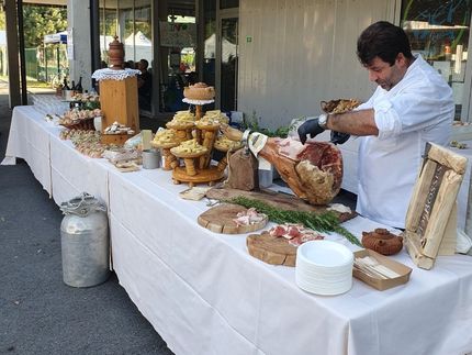 Uno chef in uniforme bianca affetta un grosso prosciutto crudo su un tavolo da buffet con pane, formaggi e salumi.