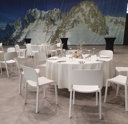 Tavoli con tovaglie bianche e sedie bianche disposti in un ampio spazio per eventi con uno sfondo raffigurante un murale di montagne innevate.