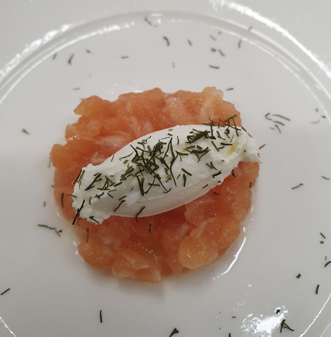 Tartare di salmone con una cucchiaiata di panna e cosparsa di aneto fresco, servita su un piatto bianco.