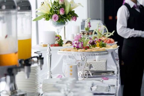 Un tavolo da buffet per catering con distributori di succhi di frutta, fiori e snack, servito da un cameriere in uniforme.