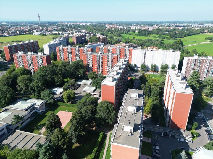 Vista aerea di diversi edifici residenziali in mattoni rossi con alberi verdi e campi, in una zona urbana in una giornata di sole.