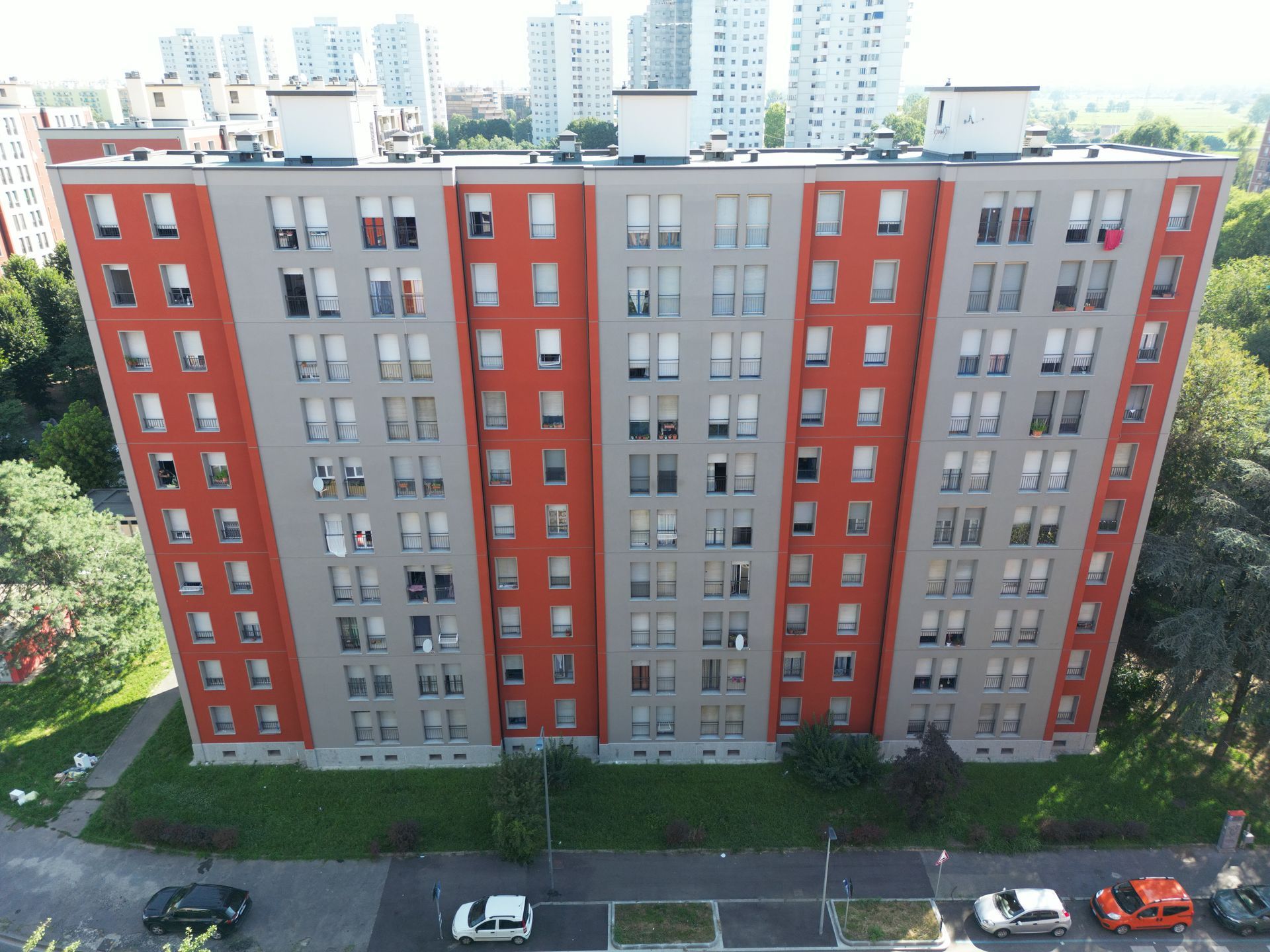Alto condominio, sezioni alternate grigie e arancioni, numerose finestre, auto parcheggiate sotto, alberi tutt'intorno.