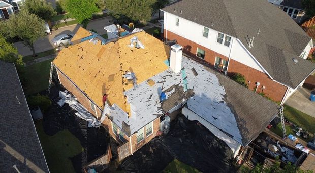 Aerial view of a house with a damaged roof and exposed plywood beside neighboring homes.