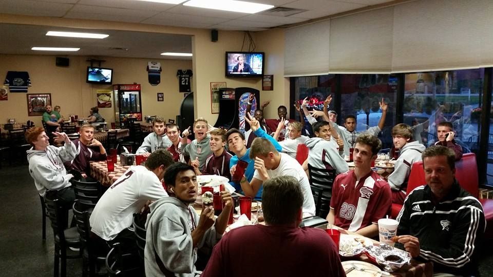 Crowded room of people seated at tables, many in red and white shirts, watching a TV screen.