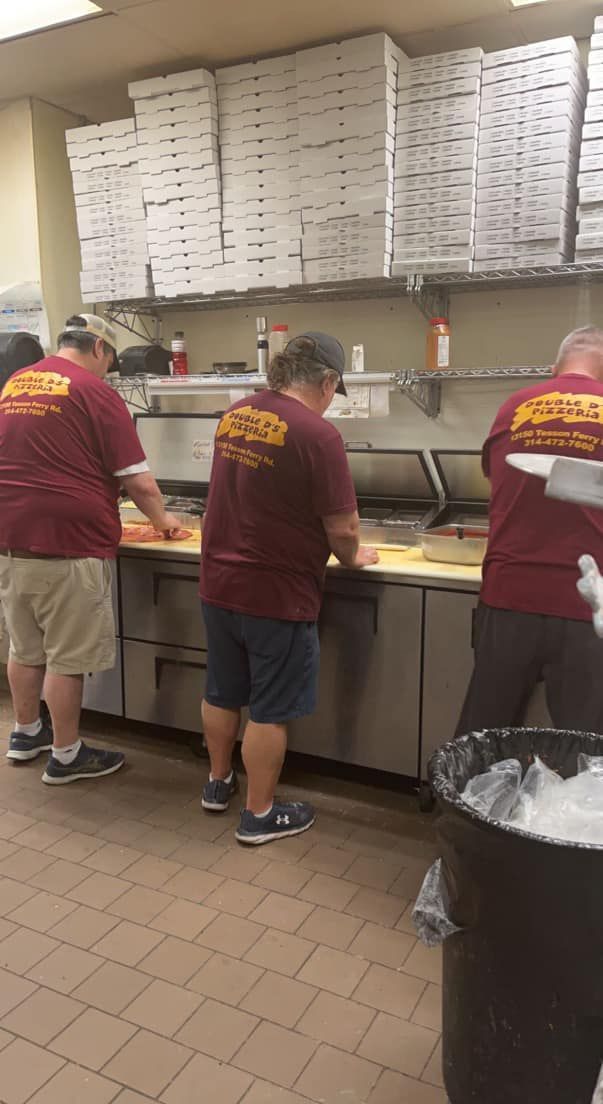 Workers in maroon shirts preparing food in a commercial kitchen with stacks of boxes overhead