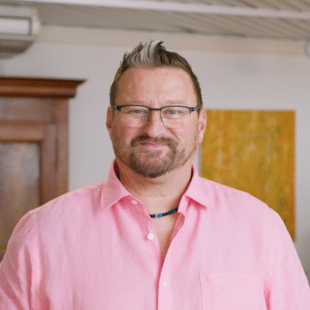 Un homme portant des lunettes et une chemise rose sourit à la caméra.