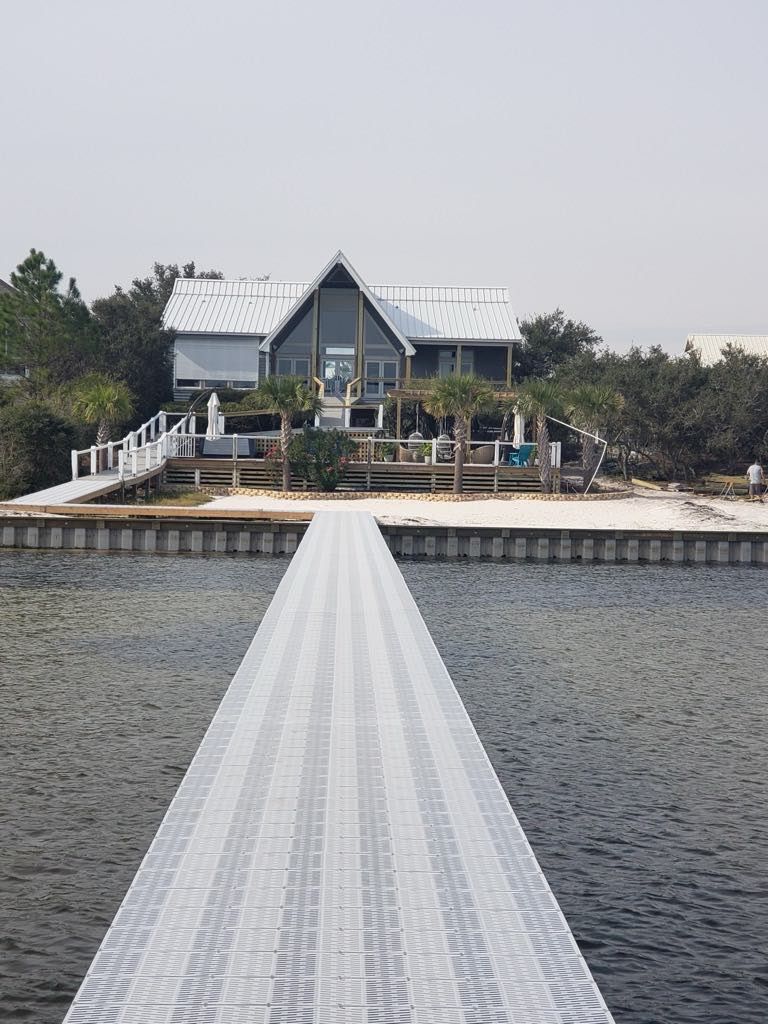 Waterfront house with a long pier extending into the water. Gray and white structure, surrounded by trees.