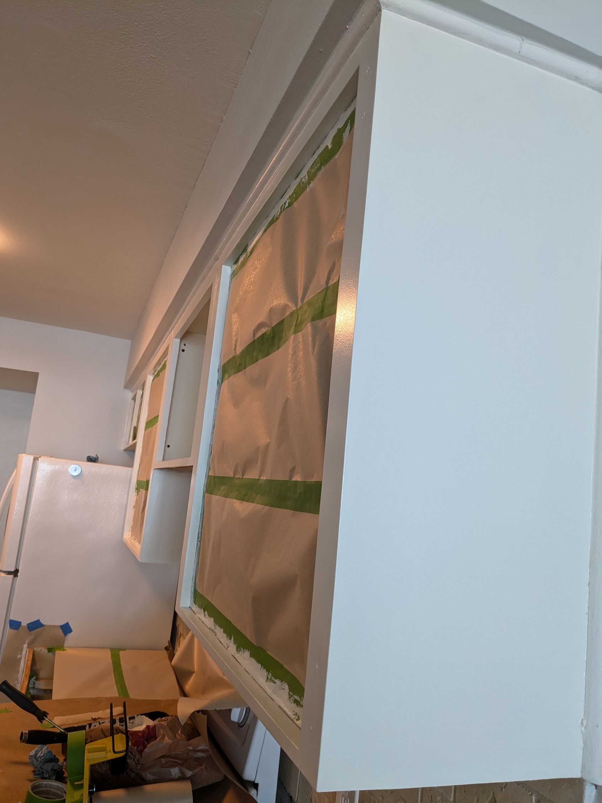White kitchen cabinets, masked and taped for painting, with visible brushstrokes.