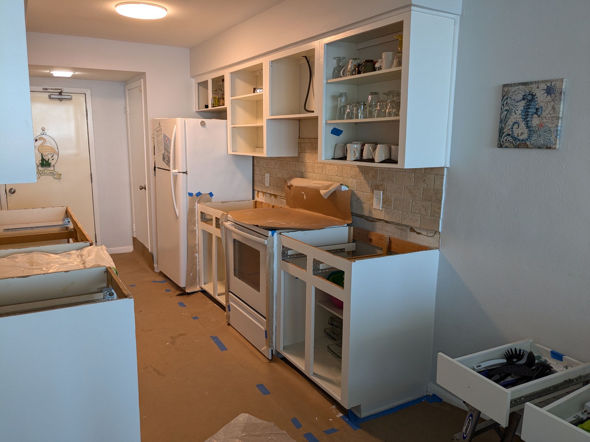 Kitchen undergoing renovation; white cabinets, appliances, and walls, with construction materials on the floor.