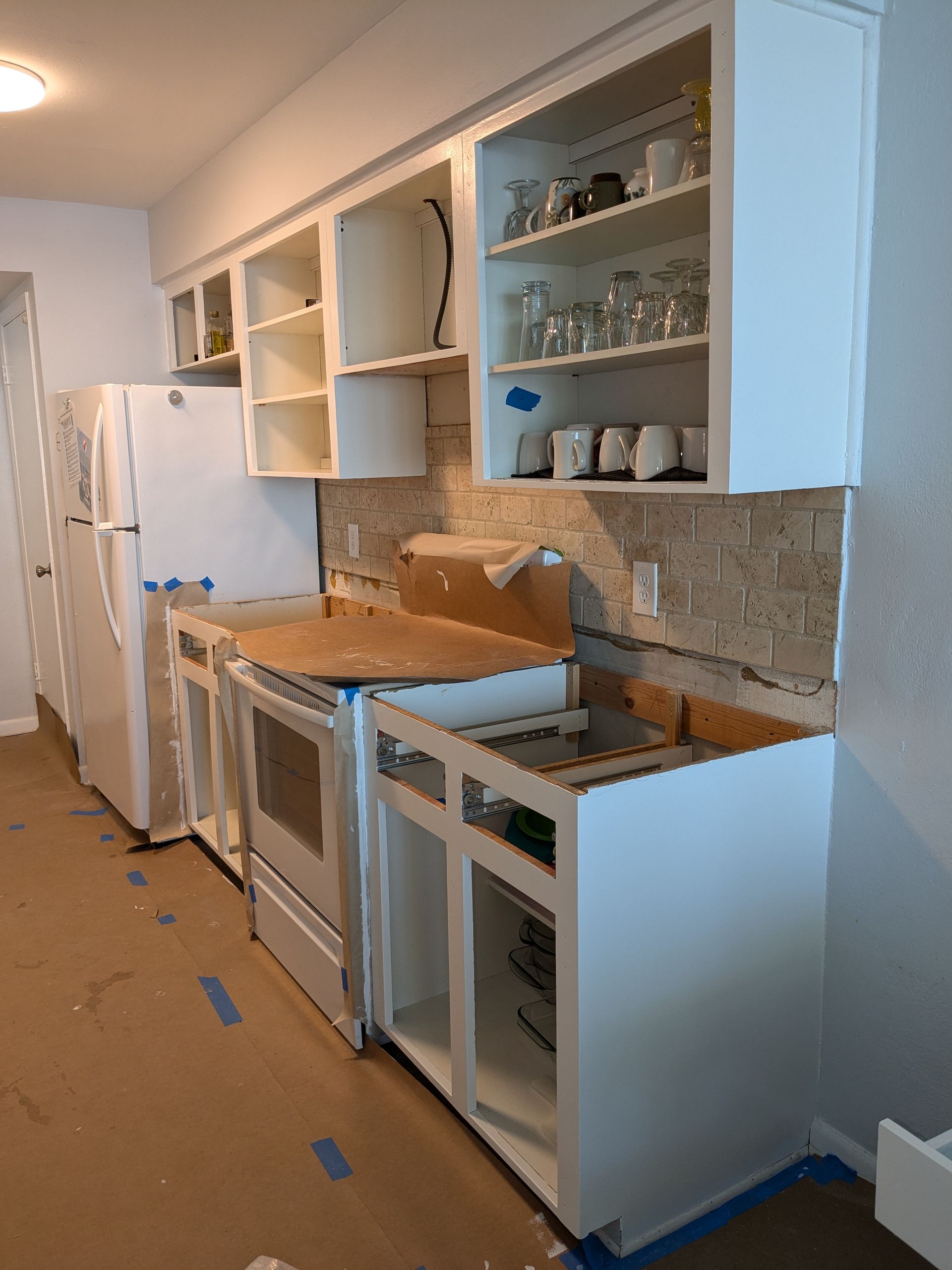 Kitchen renovation: white cabinets, appliances, and backsplash being installed. Brown paper covers the floor.