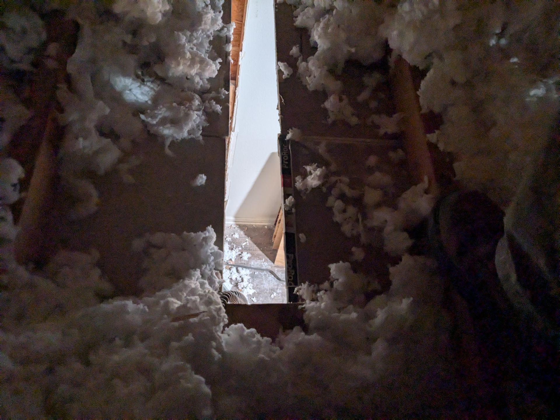 Looking up through an attic opening; fluffy white insulation surrounds the opening with light visible through the hole.