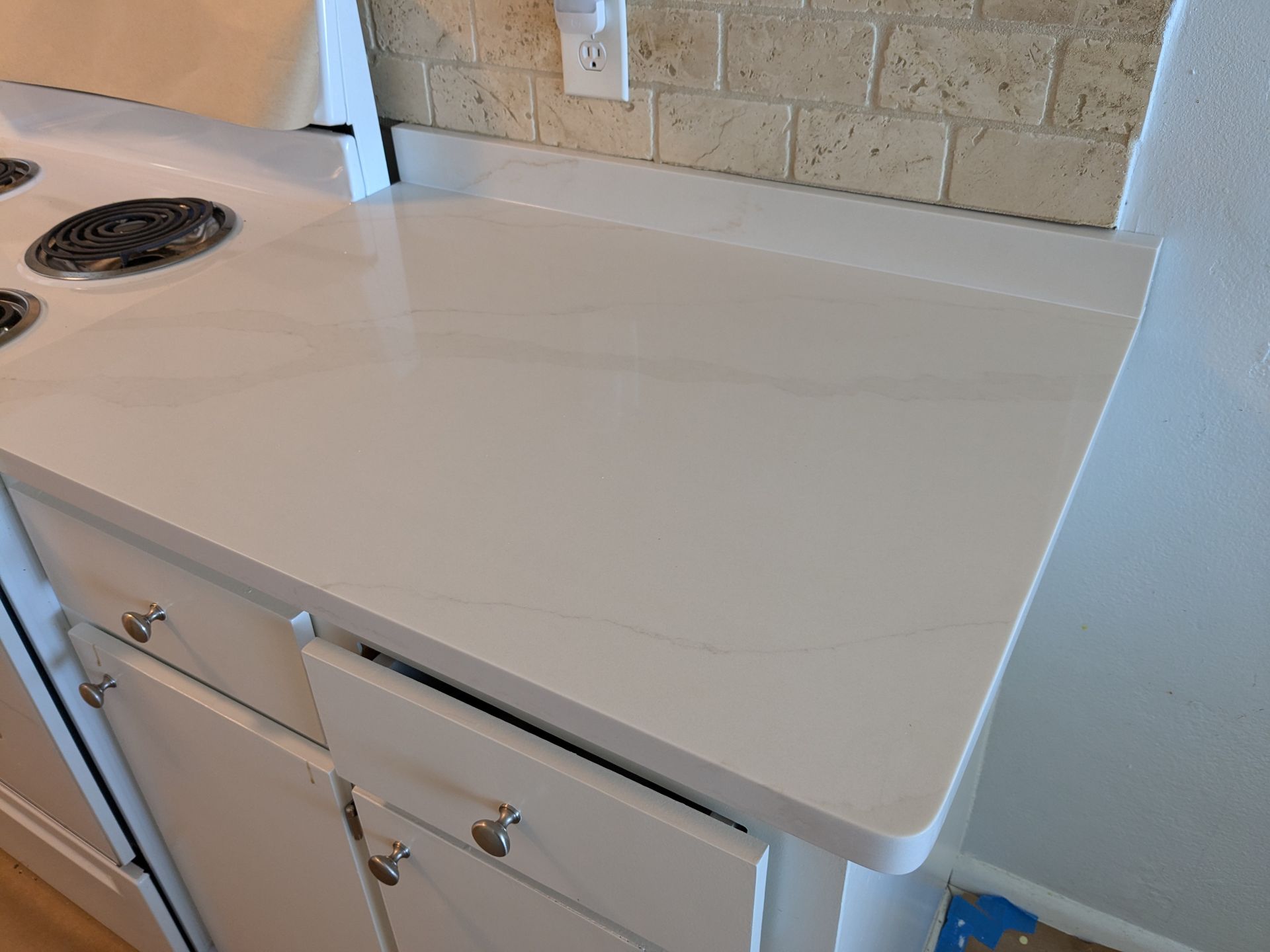 White kitchen countertop with stove and cabinets against a neutral tiled backsplash.