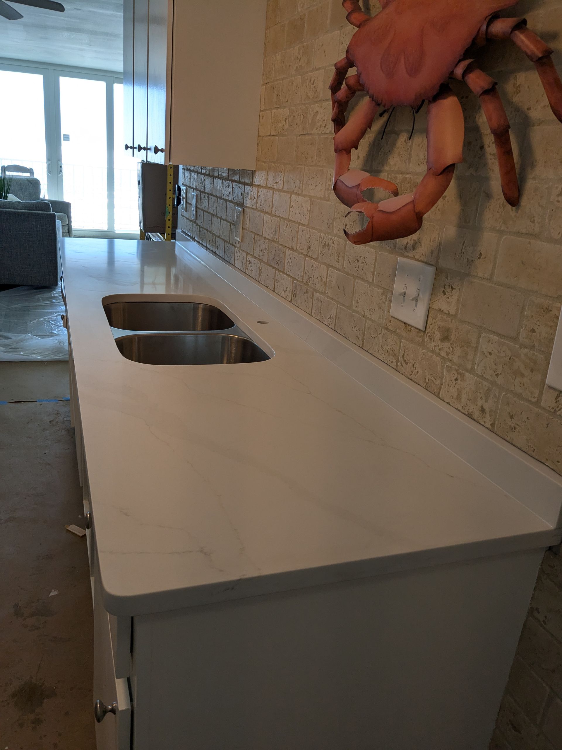 White countertop with double sink, built-in cabinet, against a brick wall, and a crab art piece.