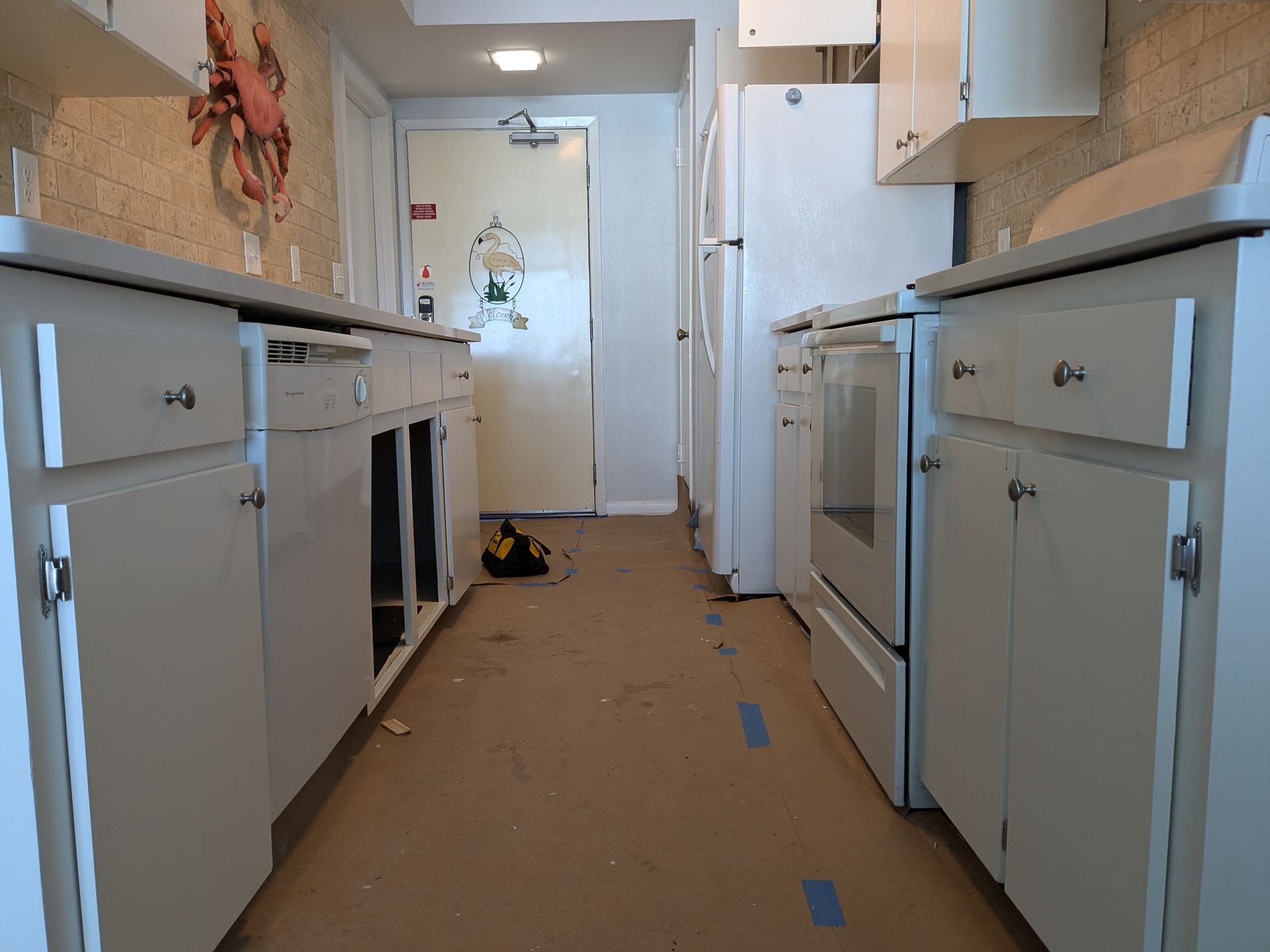Kitchen undergoing renovation; white cabinets, oven, and refrigerator line the sides, flooring exposed.