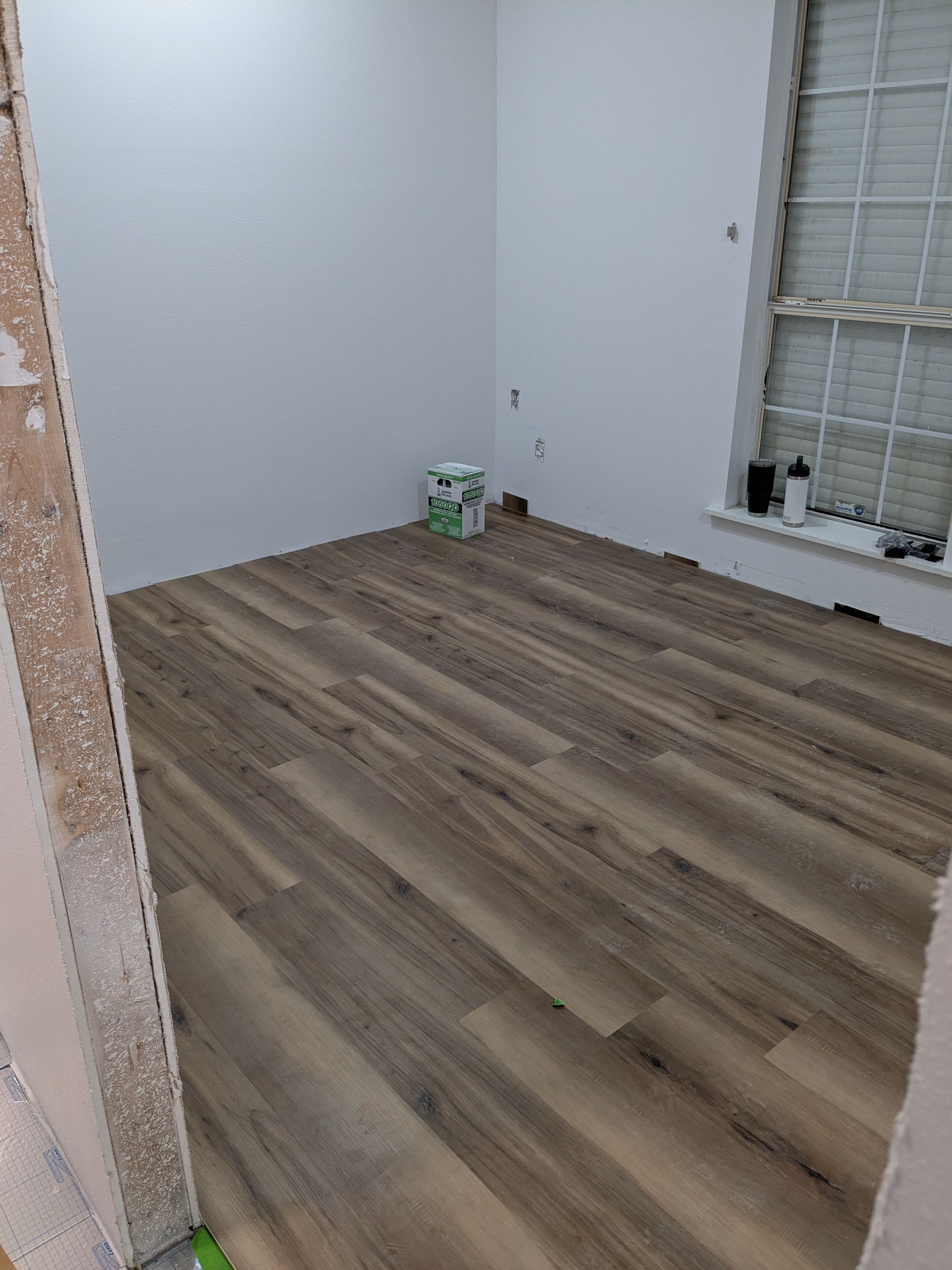 Empty room with wood-look flooring and white walls. A small container sits in the corner by a window.