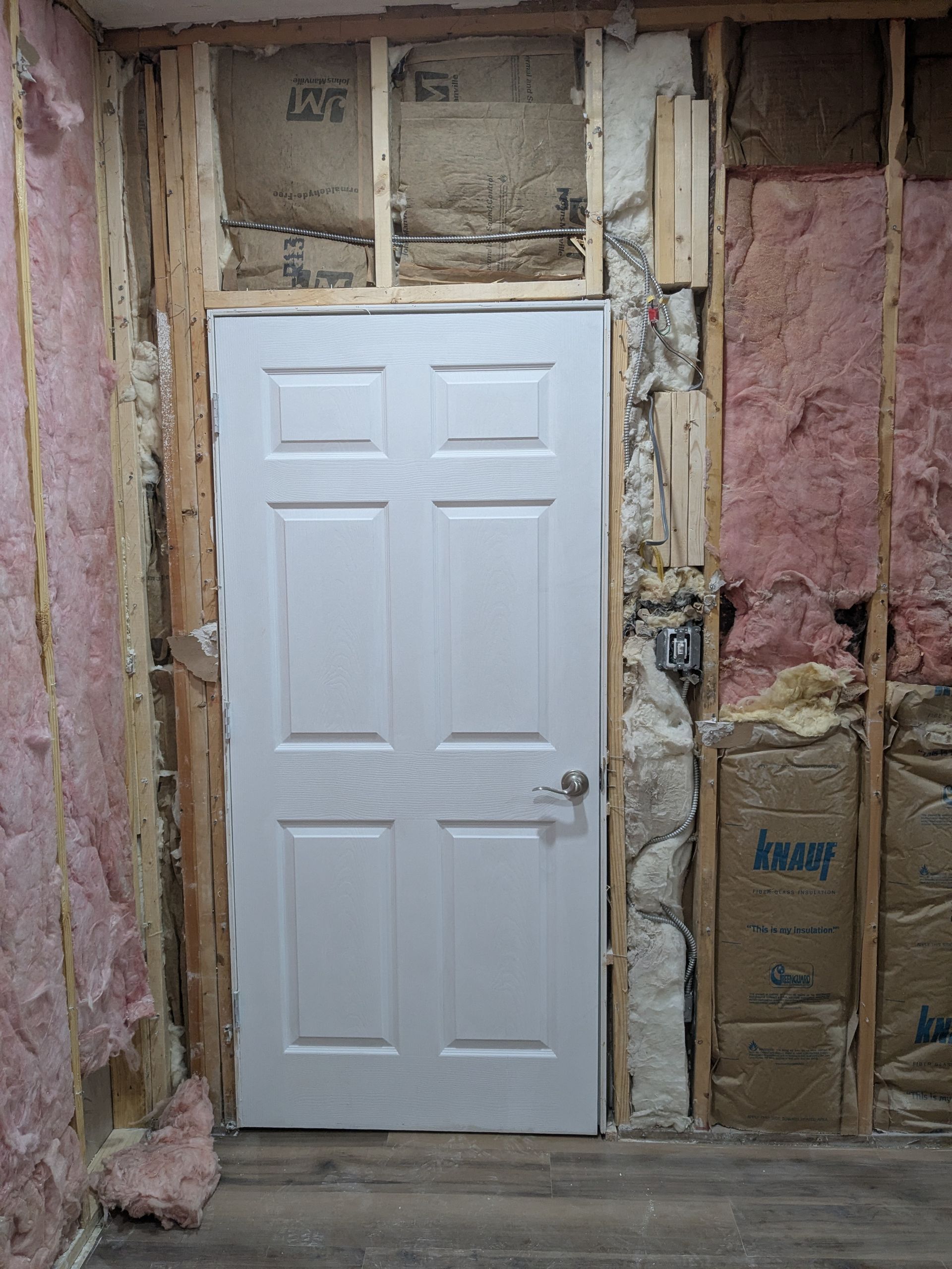 White door in a partially finished room. Exposed wall studs, insulation, and electrical box are visible.