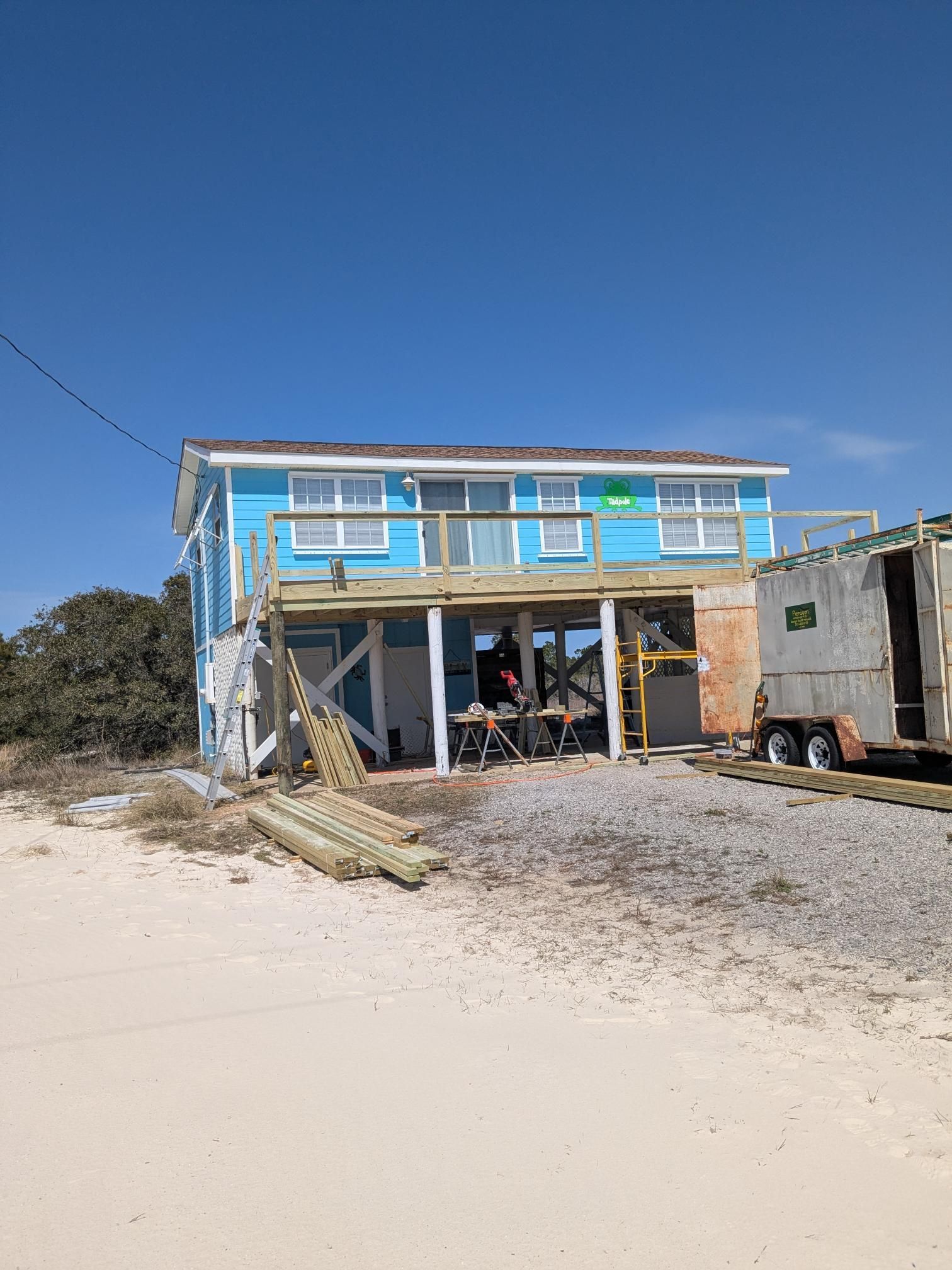 Light blue beach house on stilts with deck being built, construction materials, gravel, and sandy beach.
