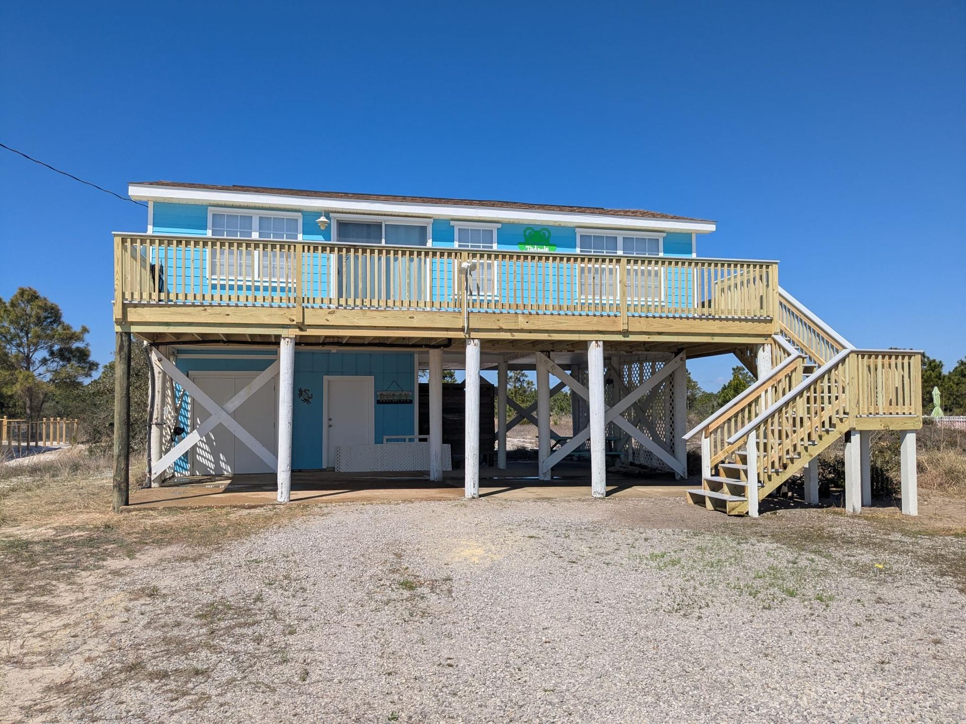 Two-story beach house, turquoise siding, wooden deck, white support beams, gravel driveway, clear blue sky.