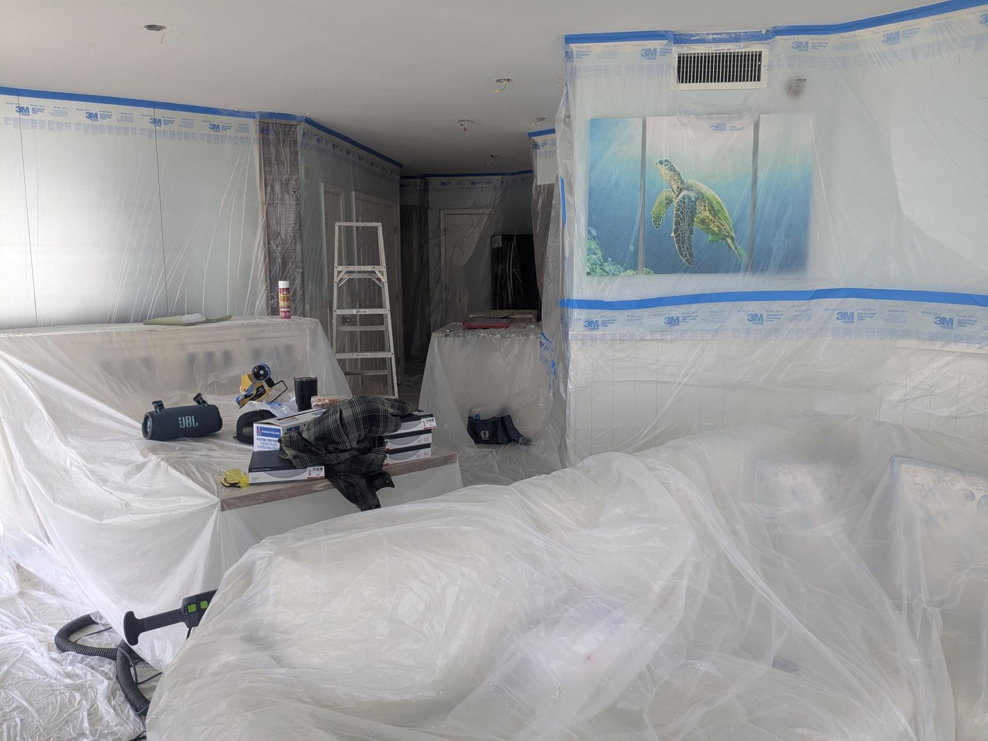 Room covered in plastic sheeting for renovation; ladder, artwork, and tools visible.
