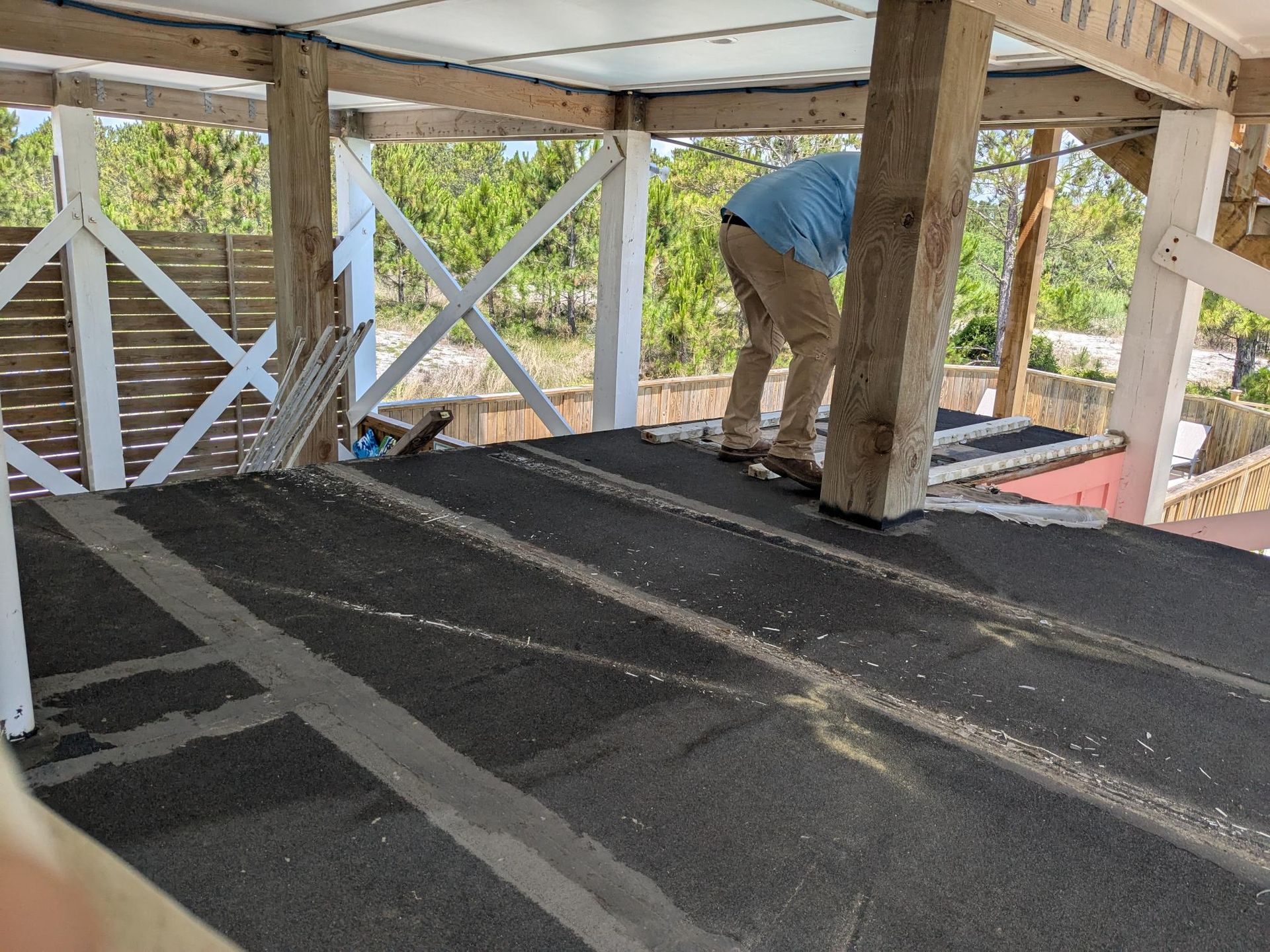 Person working on a roof, covering it with black roofing material. Wooden structure, trees in background.