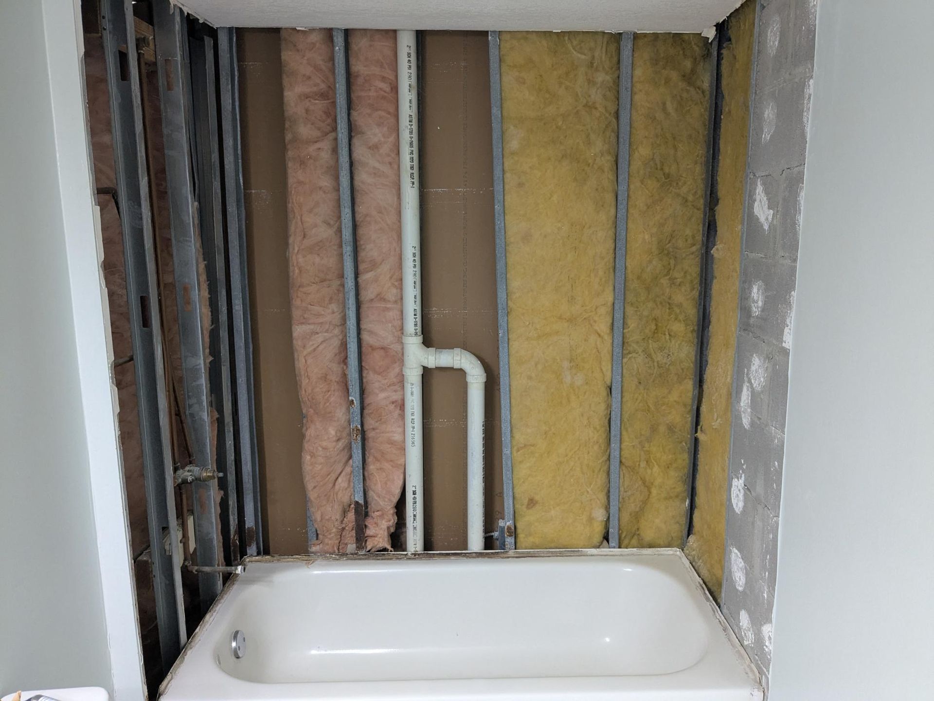 Bathroom wall under renovation, revealing plumbing, insulation, and tub. Gray studs, white tub.