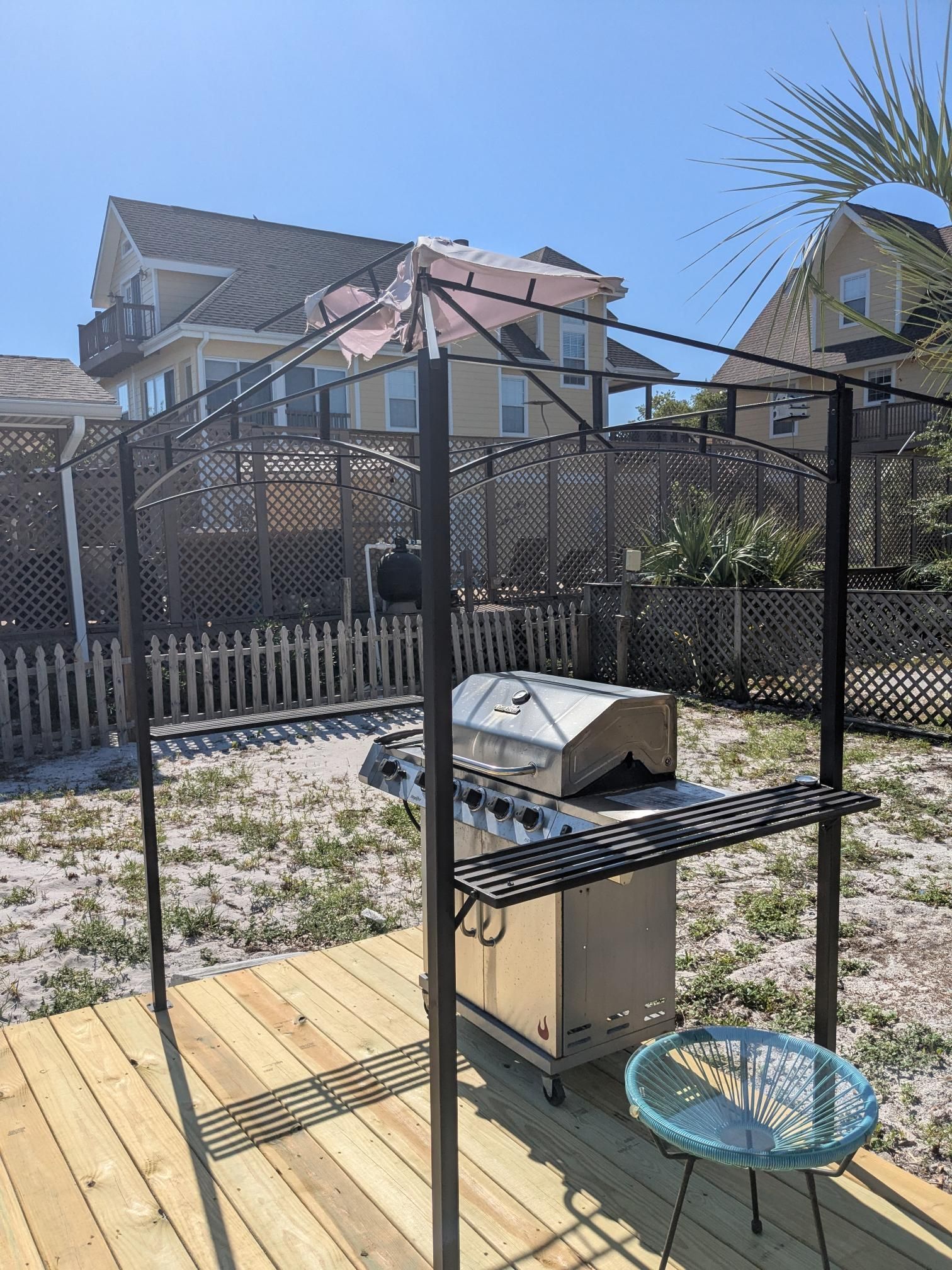 A barbecue grill with a metal frame and overhead shade on a wooden deck in a backyard.