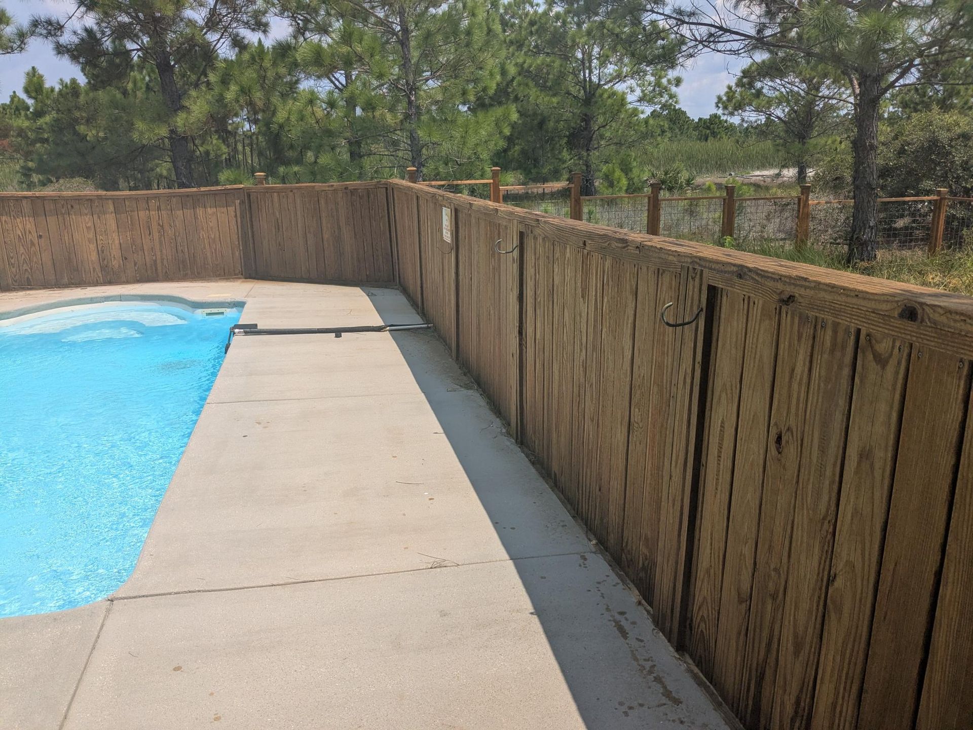 Poolside view with a rectangular pool, concrete path, and a wooden fence. Trees are in the background.