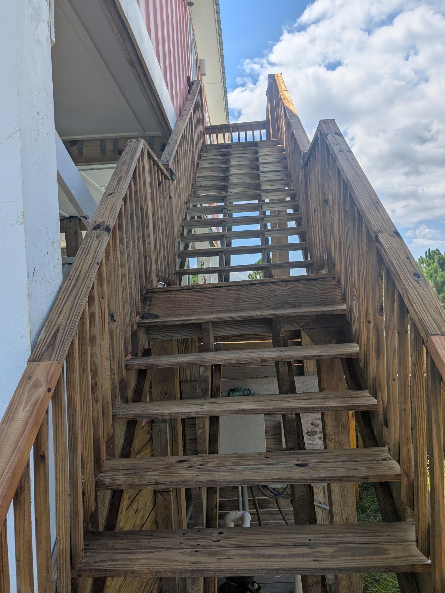 Wooden outdoor staircase leading up to a building, showing wear and tear, under a partly cloudy sky.